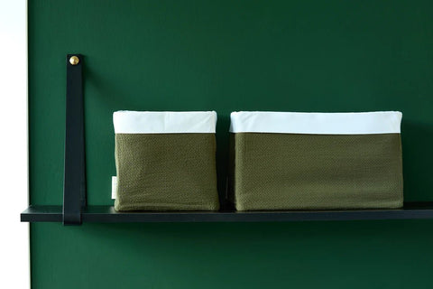 Zwei grün-weiße Stoffkörbe stehen auf einem schwarzen Wandregal vor einer dunkelgrünen Wand. Die Behälter sind unterschiedlich groß, wobei der größere auf der rechten Seite steht.