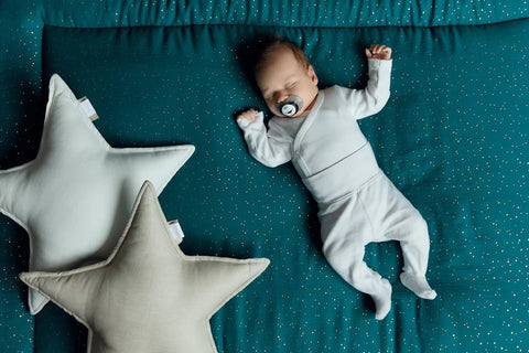 Ein Baby in einem weißen Schlafanzug schläft auf einer blaugrünen Decke mit kleinen weißen Punkten und benutzt einen Schnuller. Zwei weiche, sternförmige Kissen liegen neben dem Baby.
