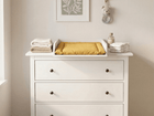 In einem minimalistischen Kinderzimmer steht eine weiße Kommode mit drei Schubladen und dem KraftKids Wickelaufsatz weiß für 108 cm breite HEMNES Kommode (Wickelfläche 50 x 70 cm), darüber eine gelbe Wickelauflage, gefaltete Handtücher und eine Flasche. Darüber hängt begrünte Kunst, durch die das Sonnenlicht fällt.