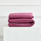 Ein ordentlich gefaltetes KraftKids Bettwäscheset Leinen magenta dunkel mit weißer Paspel, gestapelt auf einem weißen Sockel vor einer weißen Wand mit Zierleiste.