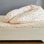 Ein sauber gemachtes Bett mit leichtem Holzrahmen ist mit cremefarbener Bettwäsche aus 100% Baumwolle mit kleinem Blumenmuster bezogen, die den KraftKids Stillkissenbezug Musselin Kirschen für Theraline 190 cm vor einer grauen Wand zeigt.