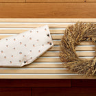 Der KraftKids Kinderwagenmuff Musselin Kirschen, gefaltet mit sichtbarem Kirschenmuster, ruht neben einem runden getrockneten Weizenkranz auf einer hellen Holzlattenbank auf einem dunkleren Holzboden und schafft eine gemütliche, einladende Atmosphäre.