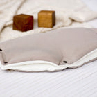 Der KraftKids Kinderwagenmuff Musselin beige, ein weicher, handgenähter rechteckiger Kinderwagenmuff aus beigem Stoff, ruht auf einem cremefarbenen Tuch mit zwei Holzklötzen im Hintergrund auf einer weiß gestreiften Fläche.