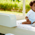 Ein kleines Kind in einem weißen Outfit sitzt lächelnd auf einem kleinen Holzbett mit dem KraftKids Bettschutzgitter Rausfallschutz mit Stauraum 60 cm breit, draußen auf einer Wiese mit grünem Laub im Hintergrund.