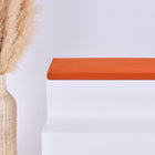 Eine hohe geflochtene Vase mit Pampasgras steht neben einem weißen Stufenpodest, auf dem das KraftKids GmbH Sitzauflage für Ikea TROFAST Doppelkrepp Rot Herbstrot Kissen liegt, alles vor einem schlichten hellen Hintergrund.