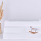 Auf einem weißen, dreistufigen Display steht die KraftKids GmbH Sitzauflage für Ikea TROFAST Musselin grau Pusteblumen neben einer beigen Vase mit Pampasgras und einem kleinen weißen Holzschaukelpferd. Der Hintergrund ist schlicht und hell.