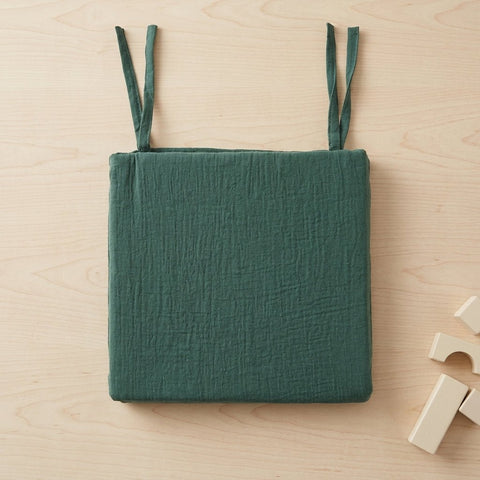 Ein grünes, quadratisches Sitzkissen für Stühle aus Musselin von KraftKids mit abnehmbarem Bezug und zwei Bindebändern liegt auf hellem Holz; unten rechts sind teils Holzbausteine zu sehen.