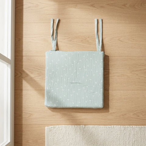 Ein KraftKids Sitzkissen für Stühle aus Musselin in Hellblau mit weißen Pusteblumenmustern und abnehmbarem Bezug ruht auf einem Holzboden neben einem cremefarbenen Teppich in der Nähe eines sonnenbeschienenen Fensters.