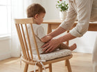 Ein Erwachsener hilft einem Kleinkind mit dem KraftKids Sitzkissen für Stühle aus Musselin in einem hellen, minimalistischen Esszimmer mit Holztisch, großem Fenster und Topfpflanze sicher auf einem gepolsterten Holzstuhl zu sitzen.