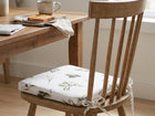 Ein Holzstuhl mit einem KraftKids Sitzkissen für Stühle aus Musselin auf der Sitzfläche sitzt neben einem Holztisch mit Tasse, Toast und Serviette in einem gemütlichen, hellen Raum. Der abnehmbare Bezug des Kissens bietet zusätzlichen Komfort und Bequemlichkeit.