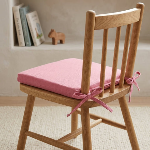 Ein Holzstuhl mit einem rosa KraftKids Sitzkissen für Stühle aus Musselin an der Rückenlehne steht auf einem hellen Teppich, während Bücher und ein kleines Holztier das Regal im Hintergrund bereichern.