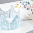 Die KraftKids Stoffkrone "weiße Pfeile auf Blau/weiße Pfeile auf Grau" liegt auf einem weißen Untergrund, während ein weicher Stern-Zauberstab und bunte Filzkugeln im Hintergrund verschwimmen-ideal für jede Kinderparty.