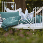 Auf einem weißen Metallbett mit grünen und gestreiften Kissen liegt das KraftKids Bettwäscheset Musselin nilblau aus 100% Baumwolle im Freien zwischen Wildblumen - perfekt für den erholsamen Schlaf unter sternförmigem Dekor und üppigem Grün.