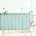 Die nackten Füße eines Babys schauen aus einer mintgrünen Bettumrandung Nestchen Musselin von KraftKids aus 100 % Baumwolle hervor, die in einem gepolsterten Stubenwagen mit Metallstäben auf einem Holzgestell vor einer hellen Wand steht.