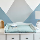 Ein moderner Wickeltisch mit der KraftKids Wickelauflage Musselin mint, gefalteter Babykleidung, Plüschtieren, einer Lampe und dekorativen Holzhausregalen steht vor einer blau-weißen, geometrischen Wand.