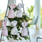 Sechs pastellfarbene Stoffbeutel aus dem KraftKids Adventskalender rosa grau hängen auf einem dreieckigen Display mit Tannenzweigen und weißen Ornamenten in einem hellen, modernen Raum mit großen Fenstern und einem verschwommenen Hintergrund.