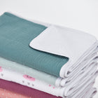 Ein Stapel pastellfarbener Babydecken und die KraftKids Wickelunterlage Musselin nilblau mit Herz- und Bärengesicht-Aufdruck sind auf einem schlichten weißen Hintergrund zu sehen.