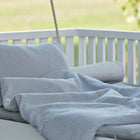 Eine gemütliche Liege im Freien mit dem KraftKids Bettwäscheset Musselin grau Punkte - ein hellgraues und weiß gepunktetes Bettwäscheset aus 100 % Baumwolle, einschließlich Kissen, Decke und Matratze - sowie einem weißen Holzgeländer und einem verschwommenen grünen Hintergrund.