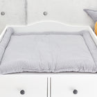 Ein weißer Wickeltisch mit der KraftKids Wickelauflage Musselin grau Punkte - mit einer gepolsterten grauen Unterlage mit kleinen weißen Punkten - steht vor einer hellgrauen Wand, dekoriert mit Stern- und Apfelkissen sowie einem Windelkorb zur Aufbewahrung.