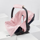 Auf einer dunklen Babyschale ist die KraftKids Decke für Babyschale Winter Musselin goldene Punkte auf Rosa drapiert, mit einer passenden rosa Griffhülle, eingebettet in weiße geometrische Blöcke vor einem minimalistischen Hintergrund.