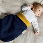 Ein Baby liegt auf einer beigen Decke, trägt ein weißes langärmeliges Oberteil und den KraftKids Strampelsack Herbst Winter Musselin dunkelblau und schaut mit erhobenem Arm nach rechts.