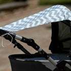 An einem grauen Kinderwagen ist der KraftKids Sonnensegel Chevron hellgrau und mint an den Griffen befestigt und bietet zusätzlichen Sitzschutz. Der Hintergrund ist im Freien und unscharf.