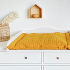 Eine weiße IDANÄS-Kommode mit einem KraftKids Wickelaufsatz, einer senfgelben Steppunterlage, einem Weidenkorb mit getrocknetem Gras und einem kleinen Holzregal zieren die weiße Wand und bilden eine stilvolle und funktionale Wickelkommode.