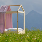 Ein einfaches Himmelbett aus Holz mit dem KraftKids Bettbaldachin Musselin goldene Punkte auf Rosa und weißem Bettzeug steht in einer grasbewachsenen Wildblumenwiese, mit Bergen im Hintergrund unter einem bewölkten Himmel.