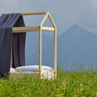 Das KraftKids Bettbaldachin Musselin goldene Punkte auf Grau ist über ein minimalistisches Holzbettgestell mit einer dachähnlichen Struktur drapiert, das auf einer Wiese mit Wildblumen, Bergen und einem bewölkten Himmel im Hintergrund steht.