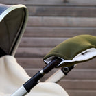 Eine Nahaufnahme eines Kinderwagens mit dem KraftKids Kinderwagenmuff Doppelkrepp Grün Herbstgrün, der am Lenker befestigt ist und die Hände warm hält, vor dem Hintergrund eines Holzzauns.