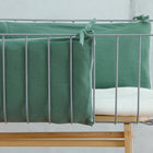 Ein modernes Babybett mit Metallrahmen bietet die KraftKids Bettumrandung Nestchen Doppelkrepp Grün Jade aus 100% Baumwolle, die an den Seiten befestigt ist, und eine weiße Matratze. Das Bettchen steht auf einem hellen Holzsockel vor einer schlichten Wand.