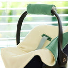 Eine Nahaufnahme einer schwarzen Babyschale im Innenbereich, bezogen mit der KraftKids Decke für Babyschale Winter Doppelkrepp Grün Jade. Das Sonnenlicht strömt durch die Jalousien und unterstreicht die gemütliche Atmosphäre und das zartgrüne Design mit Gurtschlitzen.