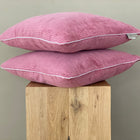 Zwei rosa KraftKids Kissenbezüge Cord Breitcord Rosa mit weißer Paspel sind auf einem hellen Holzhocker vor einem schlichten grauen Hintergrund gestapelt.