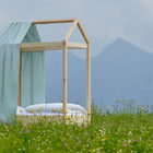 A Bettbaldachin Musselin goldene Punkte auf Grün von KraftKids schmückt ein minimalistisches Holzhausbett im Feld. Eine hellblaue Decke, Berge und Wolken sorgen für eine gemütliche Kinderzimmer-Atmosphäre.