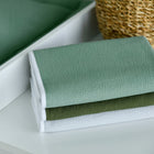 Ein Stapel von drei gefalteten strukturierten Decken in Salbeigrün, Olivgrün und Weiß ruht auf einer weißen Fläche neben einem Korb, unter dem die KraftKids Wickelunterlage Doppelkrepp Grün Jade zum zusätzlichen Schutz verstaut ist.