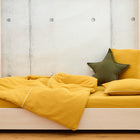 Ein Holzbett mit KraftKids Bettwäscheset Doppelkrepp Gelb Senf aus 100% Baumwolle und grünem Sternkissen, vor einer hellen Betonwand mit Kreismustern, schafft einen einladenden Raum für erholsamen Schlaf.