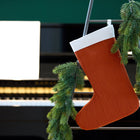 Eine KraftKids Weihnachtssocke Doppelkrepp Rot Herbstrot und eine grüne Tannengirlande hängen an einem Metallgestell vor einem Klavier mit Notenblättern und bringen festliche Weihnachtsdekoration in die Szene.