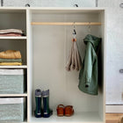 Der KraftKids Kleiderschrank für Kinder nach Montessori in Weiß bietet Regale mit gefalteter Kleidung, gemusterte Behälter, eine Stange mit einem Schal und einer grünen Jacke sowie Regenstiefel und Sandalen auf dem unteren Regal vor einer Betonwand.