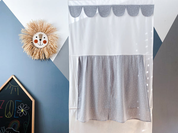 Eine dekorative Wand zeigt ein Strohlöwengesicht, eine Kreidetafel mit Zeichnungen und das KraftKids Puppentheater Kasperletheater Musselin grau Pusteblumen - ein bezaubernder Kinderbühnenvorhang - vor einem geometrisch bemalten Hintergrund.